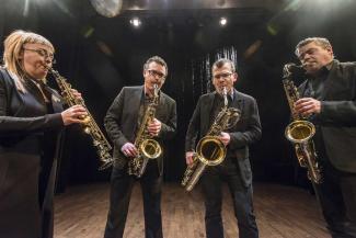 Quasar saxophone quartet Saxofonkvaretetten Quasar står i ett mörkt rum med scenbelysning och spelar på olika typer av saxofoner